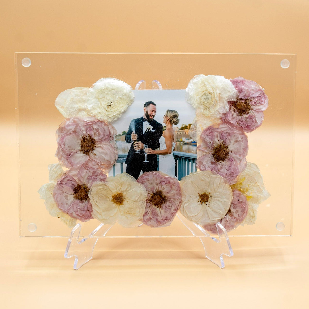 A clear resin flower preservation plate including a picture from wedding day with pressed bouquet ranunculus and dried pink roses on a clear display stand against a warm beige background.