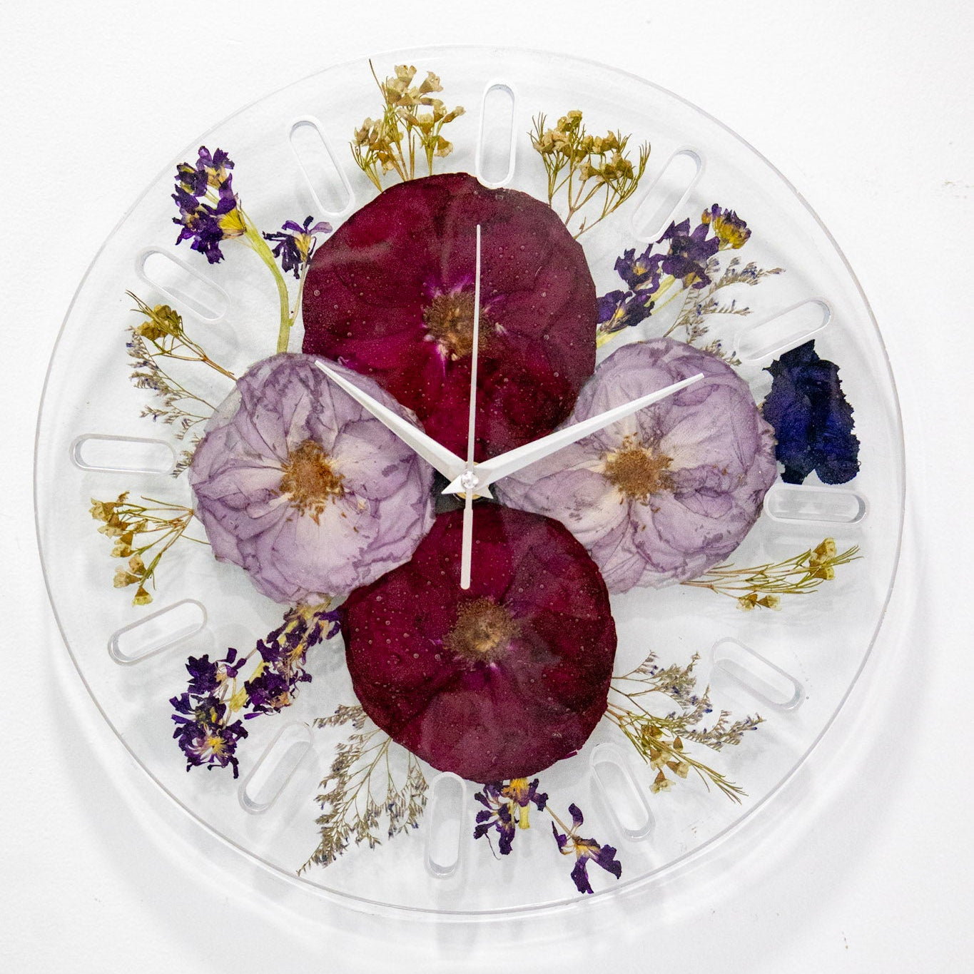 A round clear resin wall clock featuring large pressed flowers in shades of burgundy, lavender, purple, and blue, with silver clock hands, set against a plain white background.