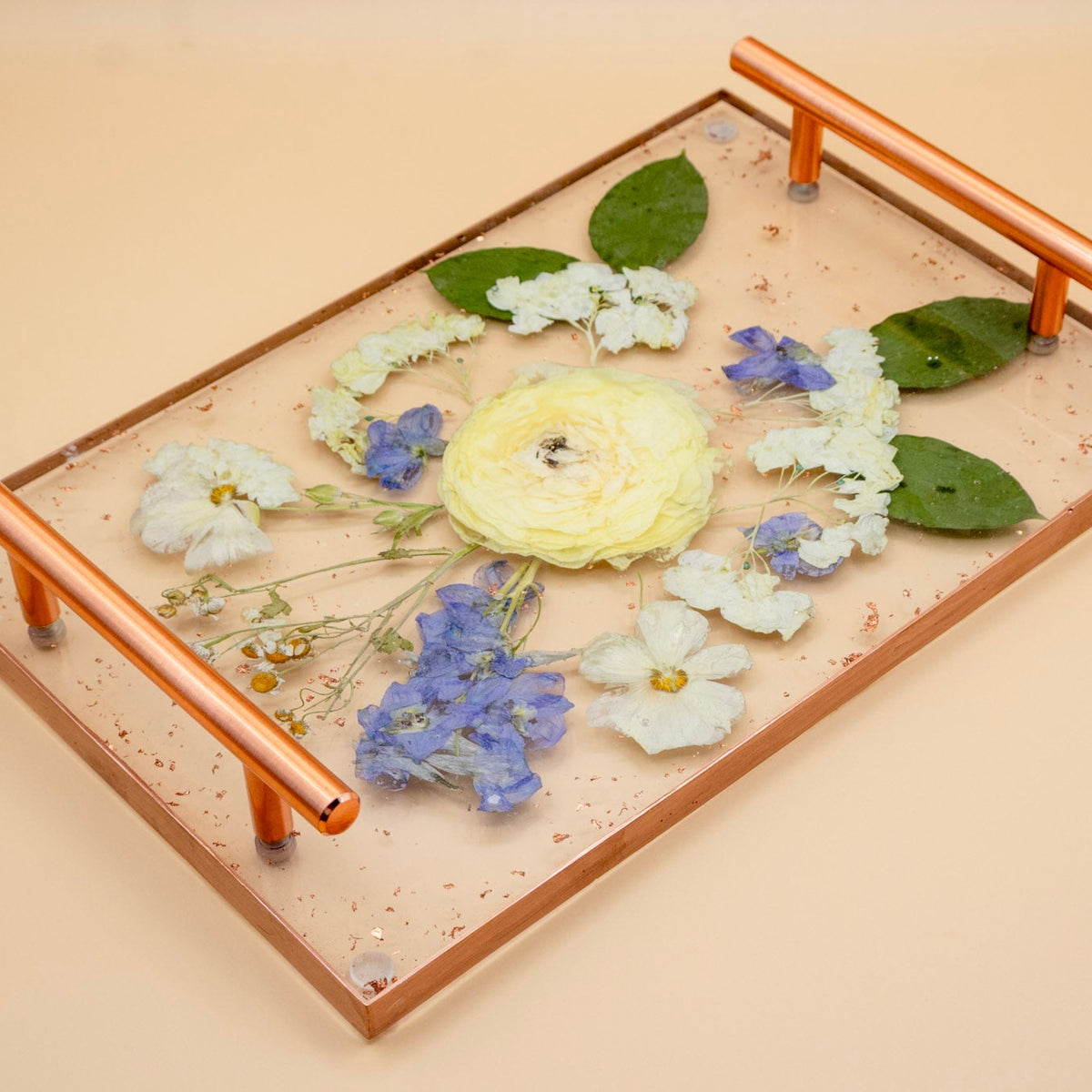 A rectangular resin flower preservation serving tray with rose gold edge and interchangeable copper handles, filled with pressed ranunculus, blue delphinium, hydrangea, butterfly ranunculus and green leaves, photographed on a warm beige background.