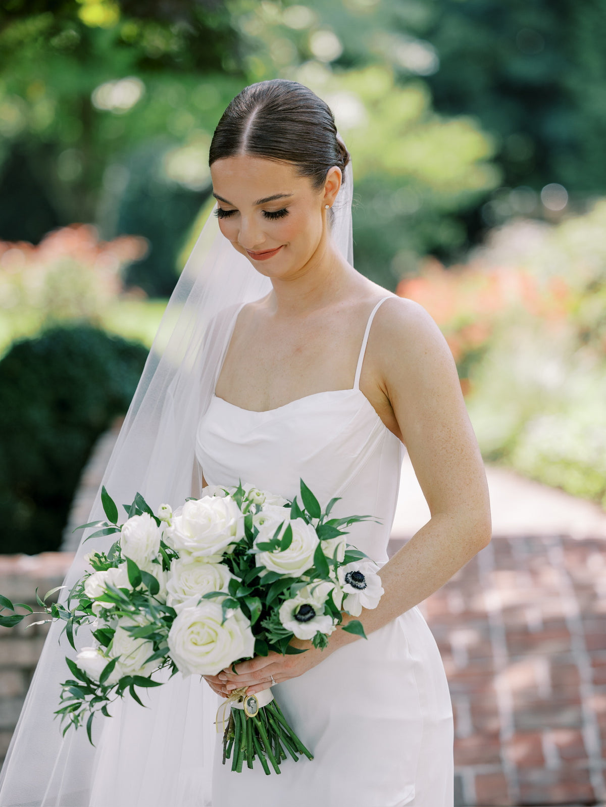 A bride in a white spaghetti-strap wedding dress and veil holds a bouquet of white roses and anemones in an outdoor garden setting.