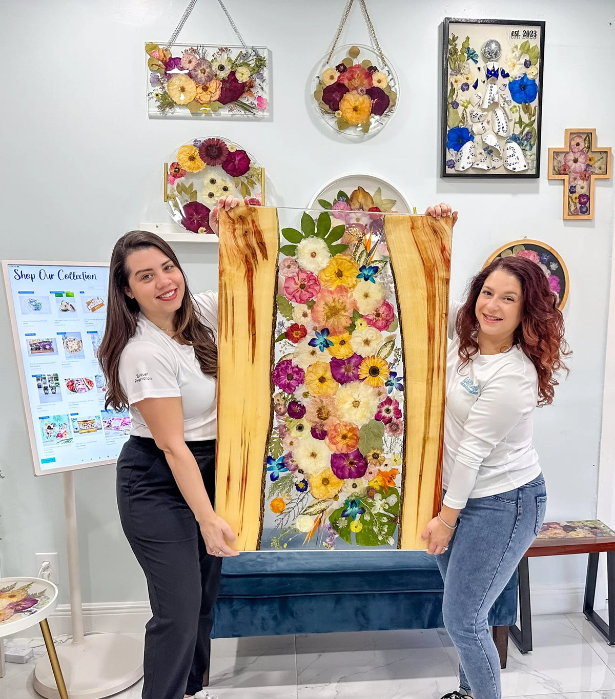 Two women holding a floral artwork in a room with decorative elements.