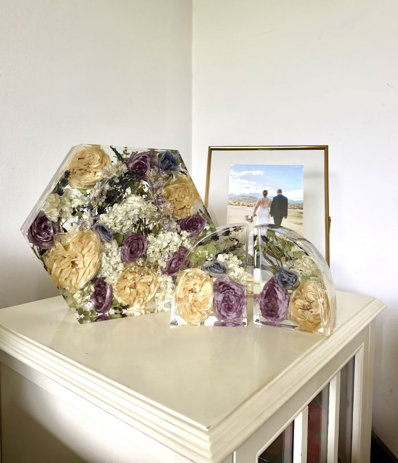 Hexagonal preserved flower arrangement in a clear container on a white surface with a framed picture in the background.